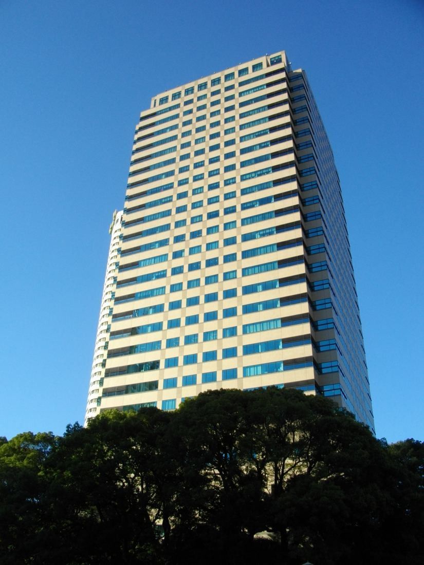 Okamura: Tall office tower with a pale stone facade against a blue sky, trees in front
