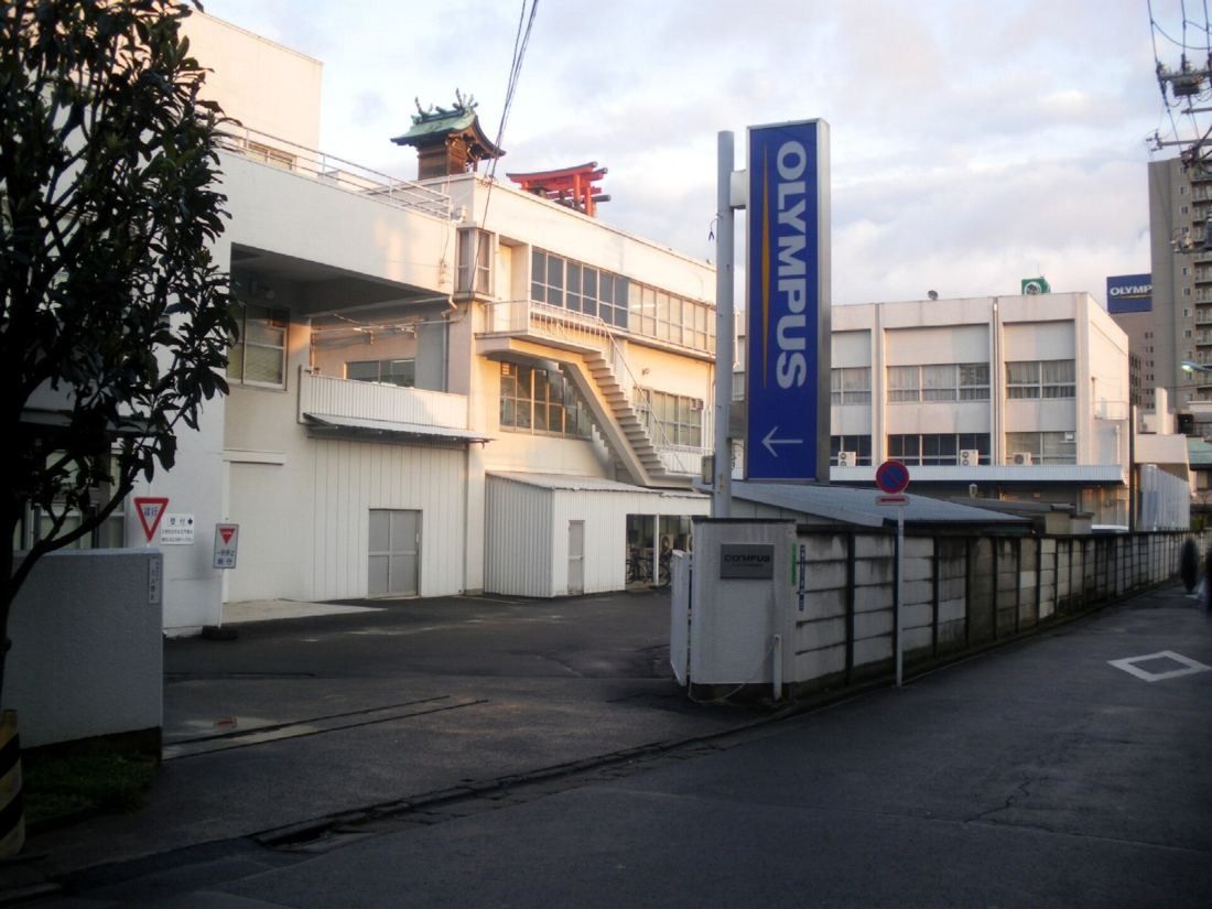 Low white factory buildings behind a wall, an Olympus sign and a shrine roof with a red torii on top