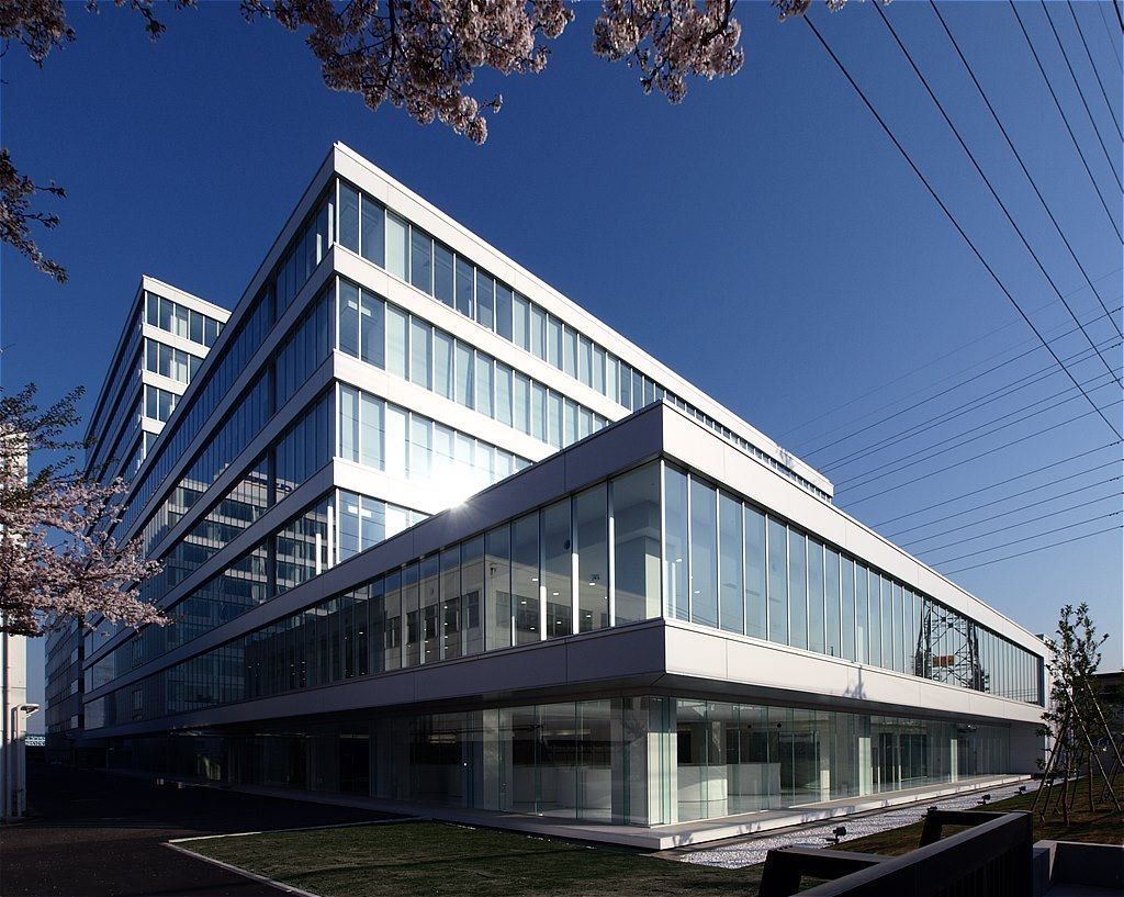Olympus: White office building with long horizontal glass bands under a blue sky, cherry blossom at the edge