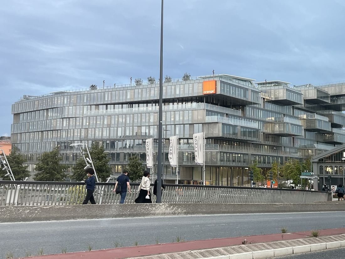 Orange: Long glass office building seen across a road and a bridge railing, with pedestrians in the distance