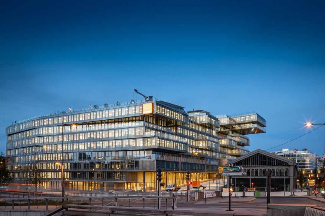 Orange: Office building with stacked floors at dusk beside an old covered market hall with a pitched roof