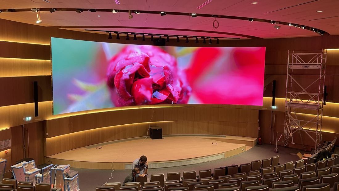 Orange: Wood-panelled auditorium with tiered seating facing a wide curved LED screen