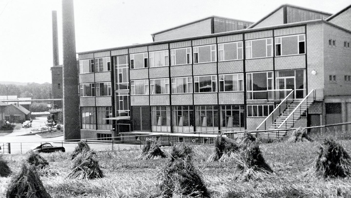 Black-and-white photo of a four-storey factory building with a tall chimney, haystacks in the field in front