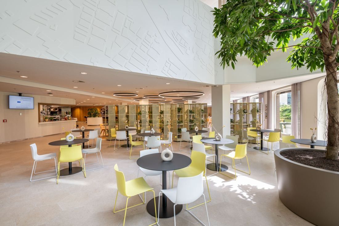 Rabobank: Bright ground-floor café with round tables, yellow and white chairs, a large tree in a planter and screens on the wall