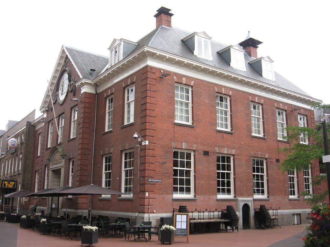 Red-brick building with a gabled front, white cornice and many tall sash windows, with café terrace tables in front