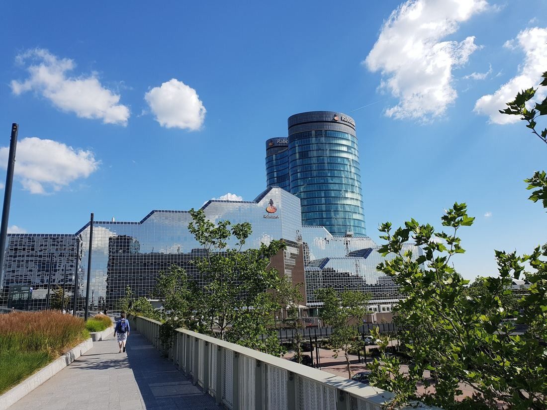 Two curved glass towers rising behind a long glass-clad building with a jagged roofline, seen from a landscaped path under a blue sky