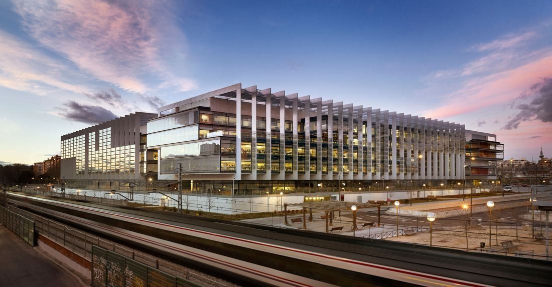 Long low glass office building with white vertical fins at dusk, railway tracks in the foreground