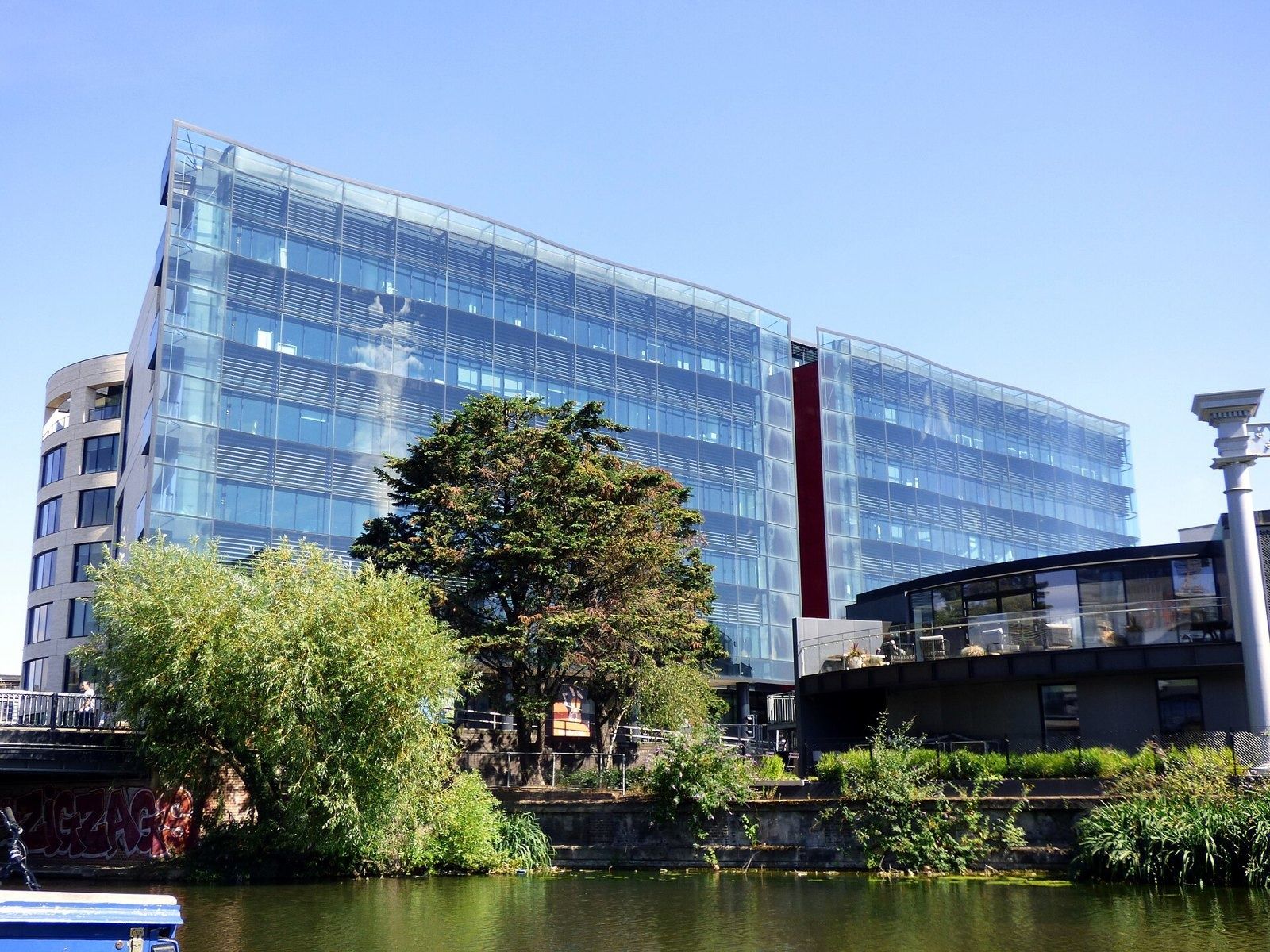Glass-fronted office building seen across the canal with trees on the bank
