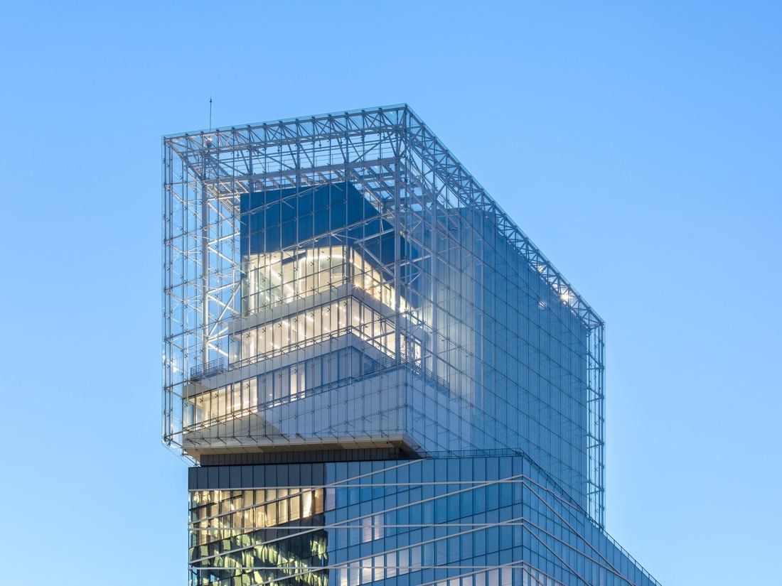 Glass crown at the top of a skyscraper against a clear blue sky, with lit floors visible inside