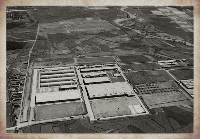 Black-and-white aerial view of long factory buildings surrounded by open farmland