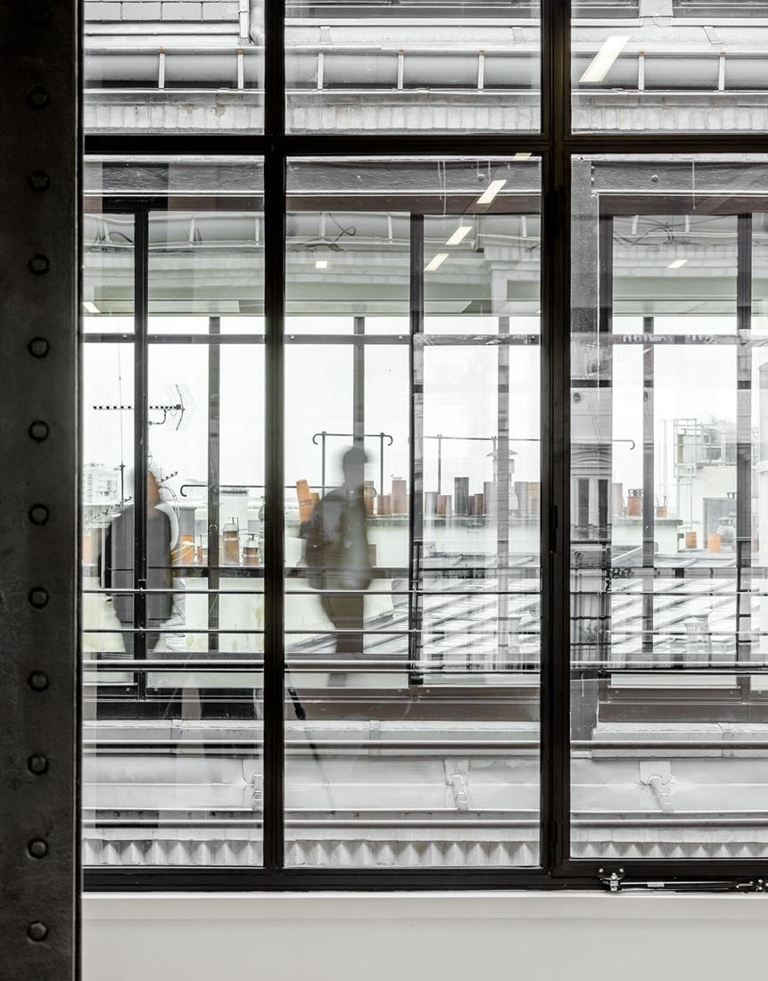 Sanofi: Riveted black steel post beside a large steel-framed window, blurred figures walking past rooftops outside