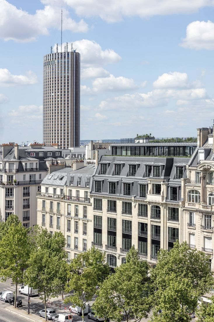 Sanofi: Stone apartment-style façades with dormer roofs and a glazed rooftop storey, a tall cylindrical tower behind