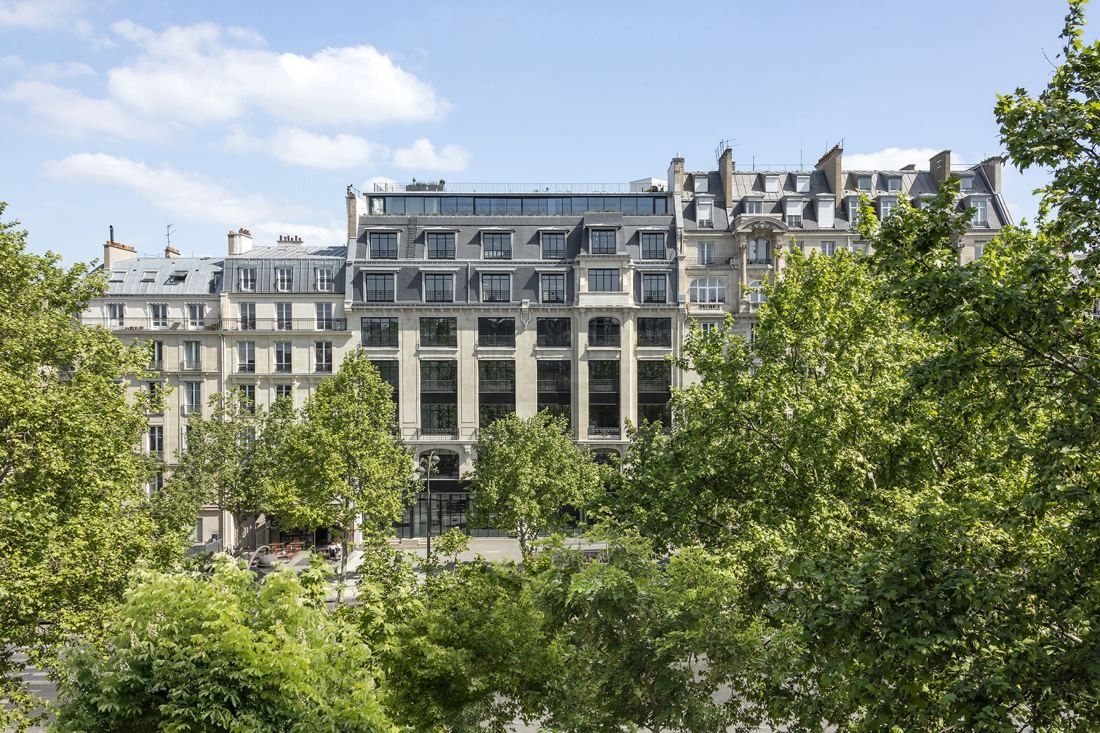 Stone Haussmann-era façade with tall black-framed windows and a glass rooftop storey, seen across tree tops on a sunny day