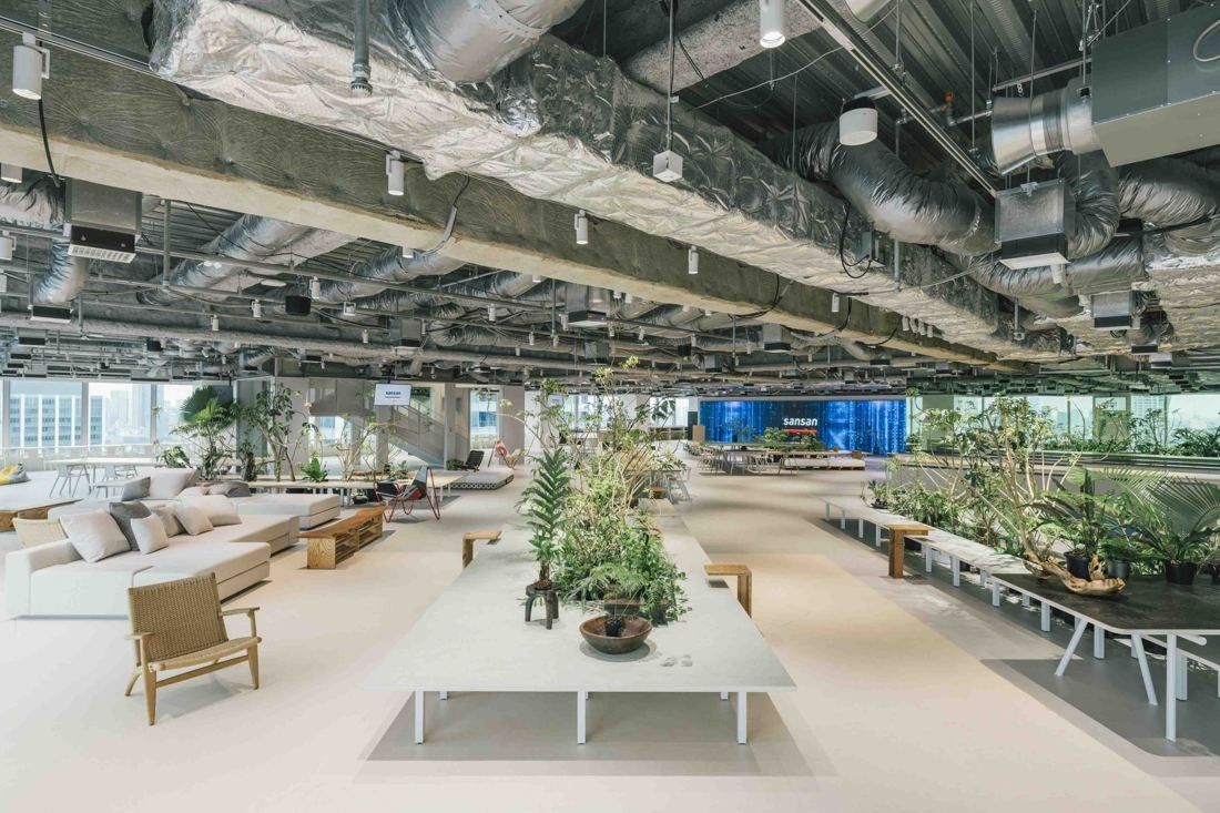 Sansan: Long open floor with exposed ducts, white tables, sofas and planters of green plants