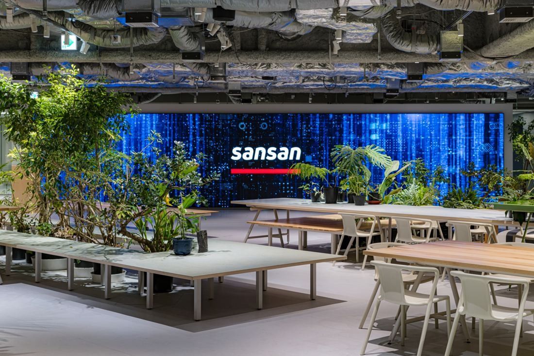 Wide open floor with long white tables, planters of tropical plants and a blue digital wall with the Sansan logo