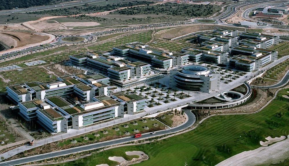 Aerial view of a campus of low white office buildings with green roofs, surrounded by lawns and roads