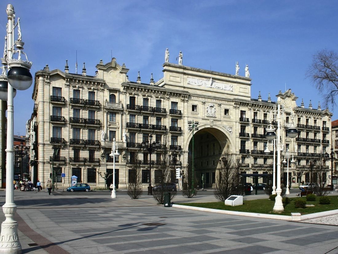 Ornate stone building with a large central arch over the street, facing a wide paved promenade