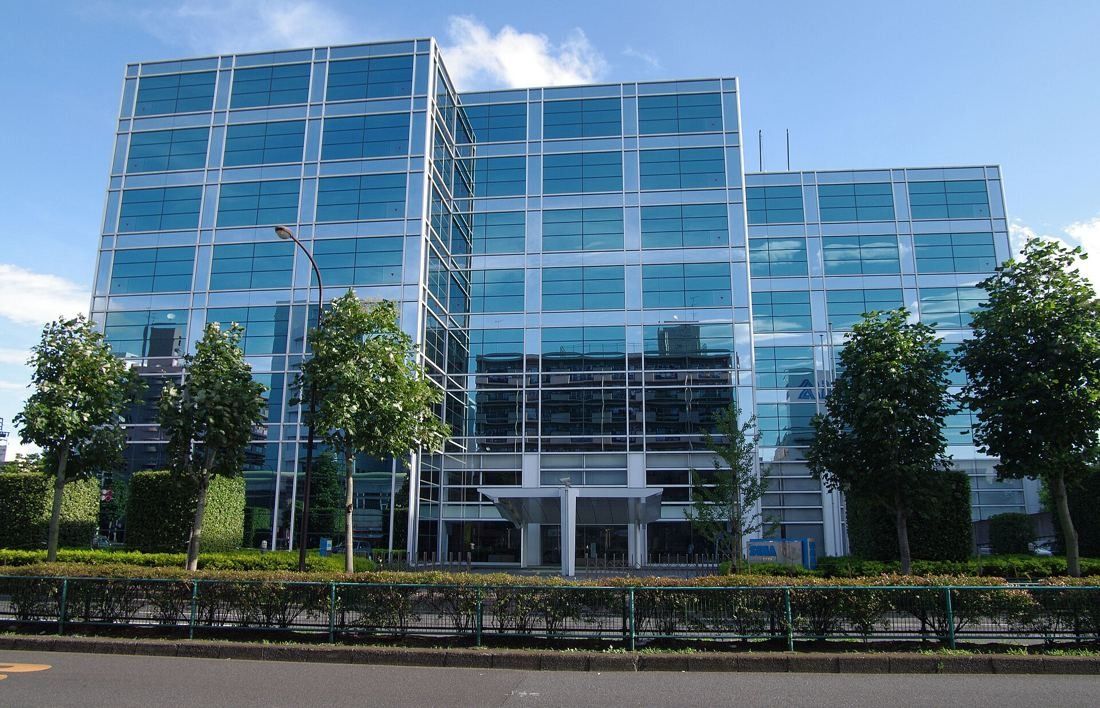 Glass-fronted office building behind a row of trees and a hedge