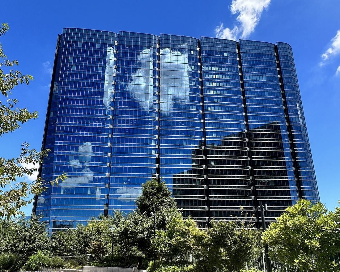 Glass office tower reflecting blue sky and clouds, with trees in front