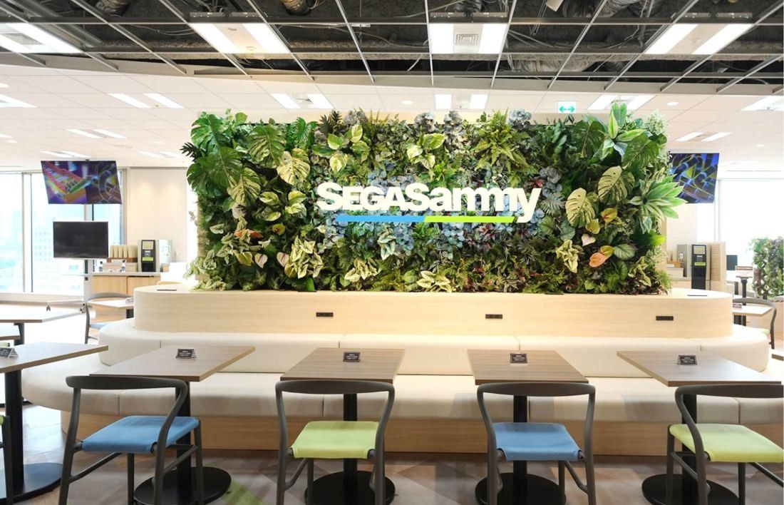 SEGA: Green plant wall with SEGA SAMMY lettering above bench seating and stools