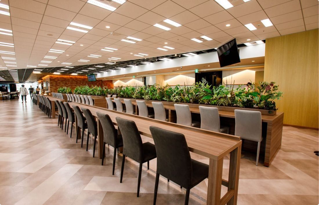 SEGA: Long communal table with dark chairs and a planter divider in a canteen