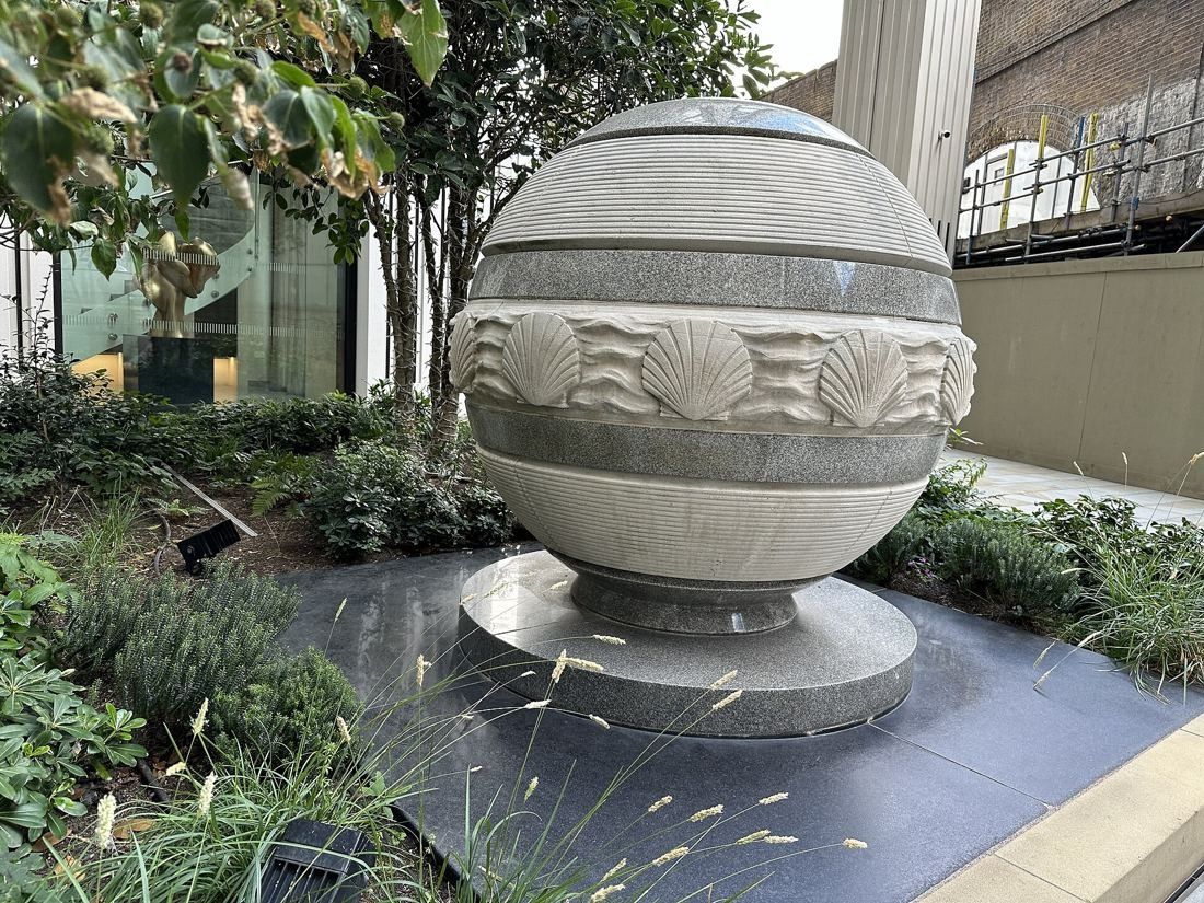 Shell: Carved stone sphere decorated with a band of scallop shells, set on a plinth among plants