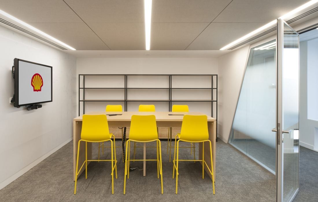 Shell: Glass-walled meeting room with bright yellow stools around a high table and a wall screen