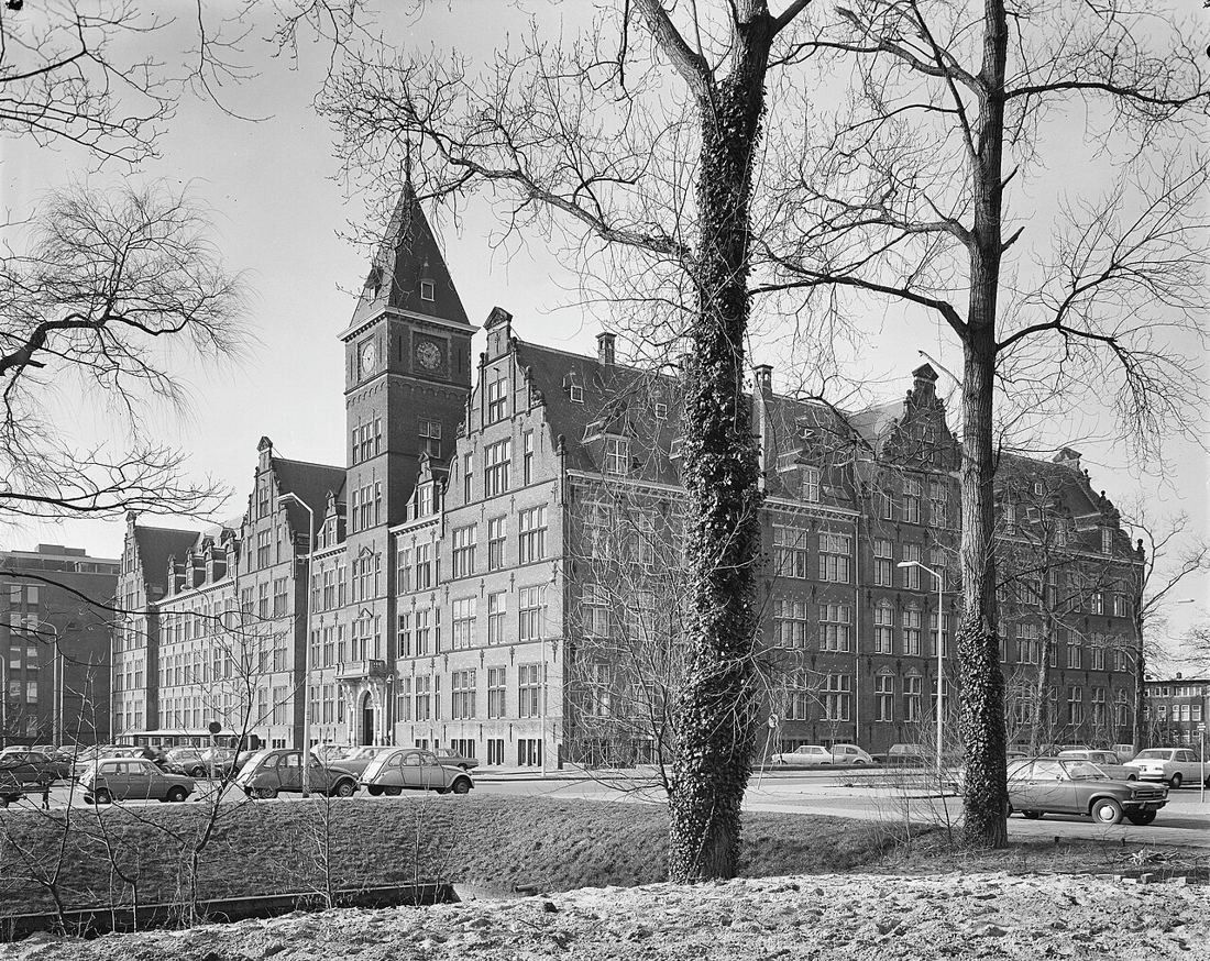 Large brick office building with gables and a clock tower behind bare trees, cars parked in front, black and white