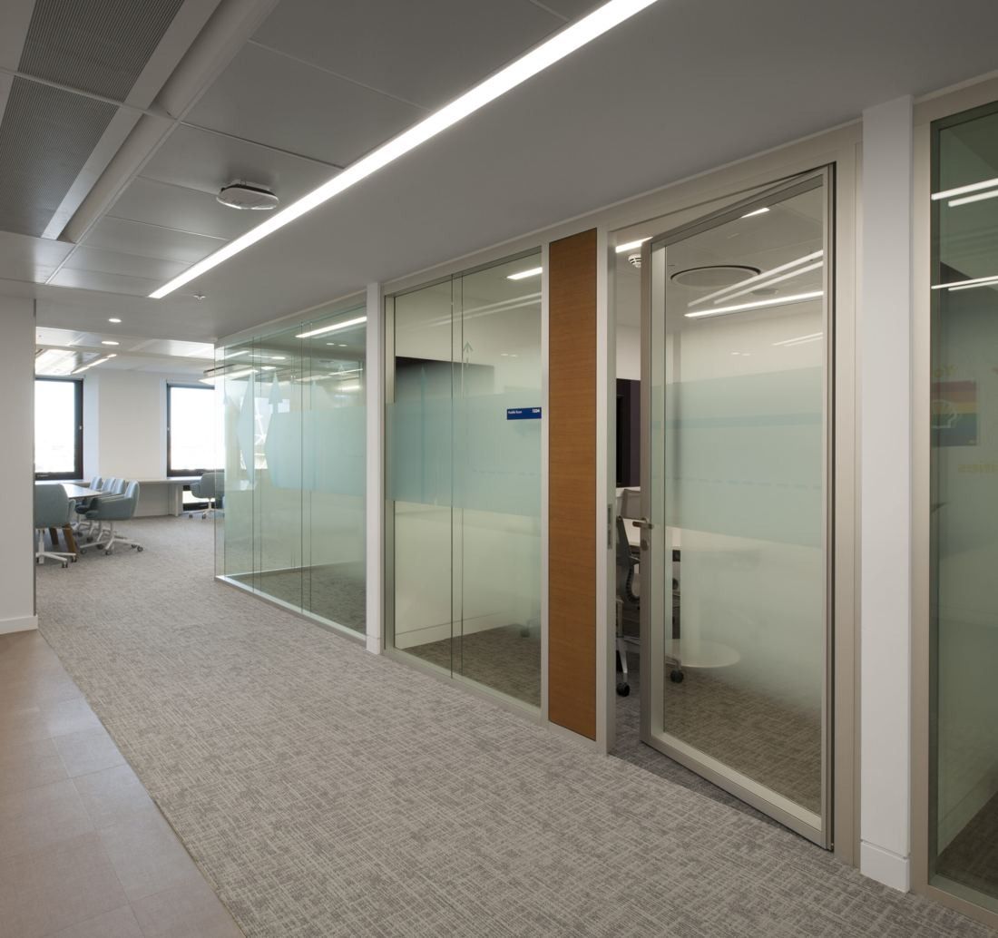 Shell: Office corridor lined with glass-walled meeting rooms and wood door panels, leading to a table area by the windows