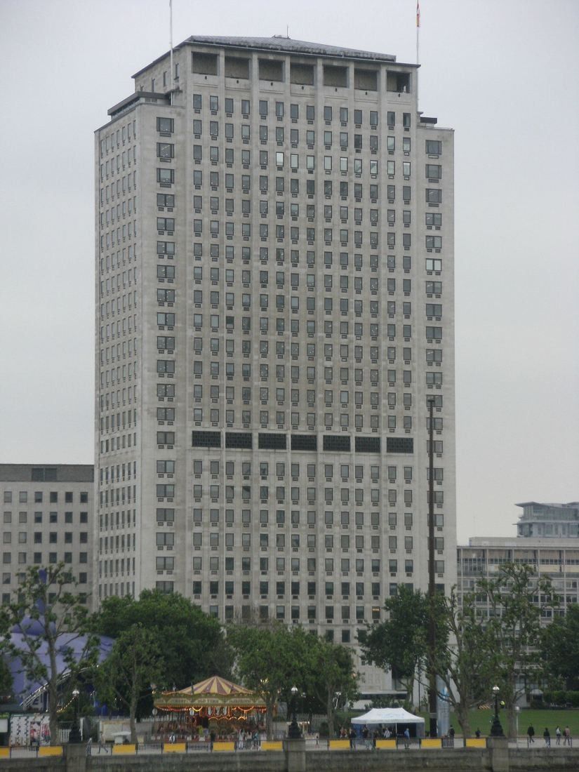 Shell: Tall stone office tower behind trees and a small carousel on a grey day