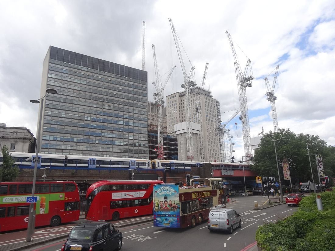 Tall stone tower surrounded by tower cranes and new buildings under construction, with buses on the road in front