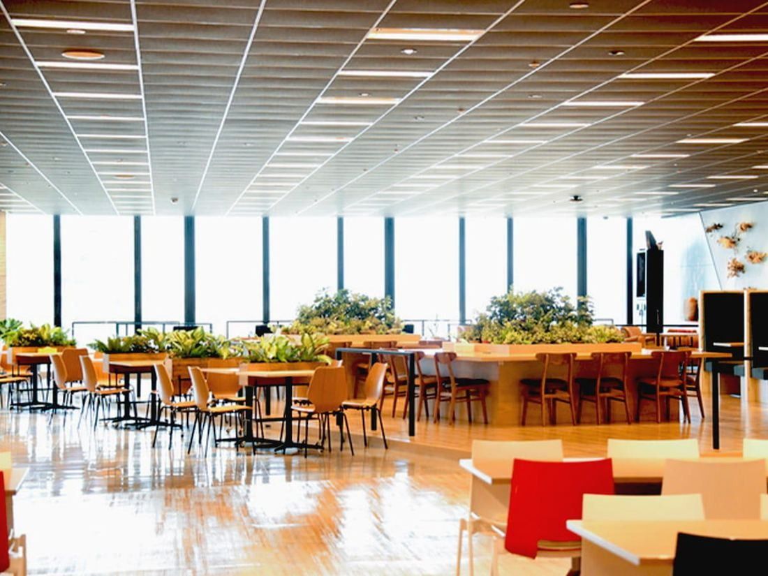 Square Enix: Bright lounge with wooden floor, rows of tables and chairs, and plants by tall windows