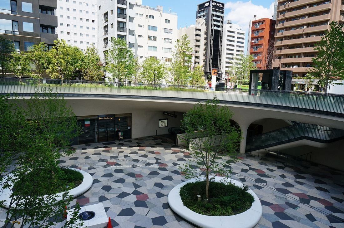 Square Enix: Sunken courtyard with patterned paving and round tree planters, apartment blocks behind