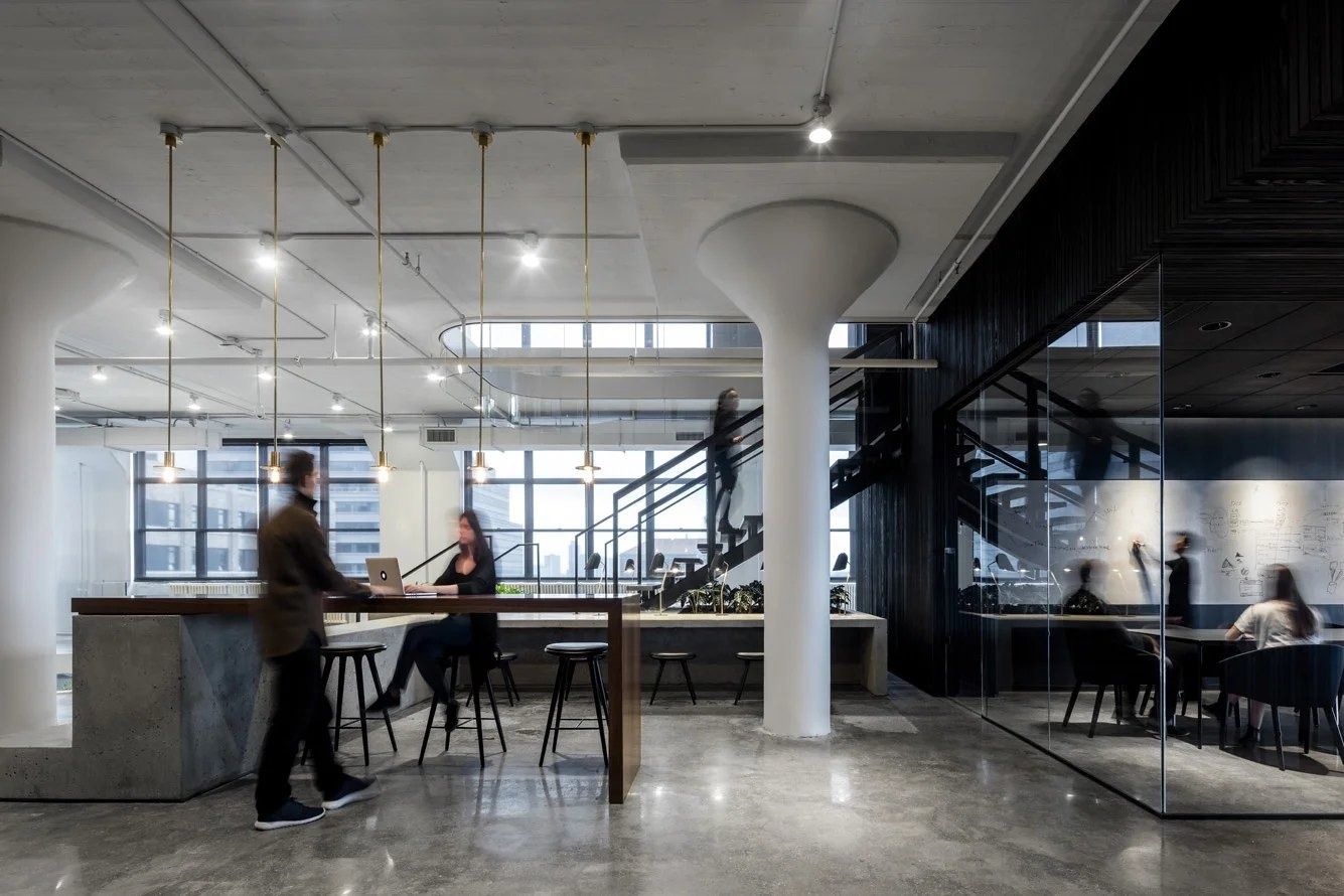 Squarespace: Concrete counter with stools and brass pendant lights, a black stair and a glass-walled meeting room behind