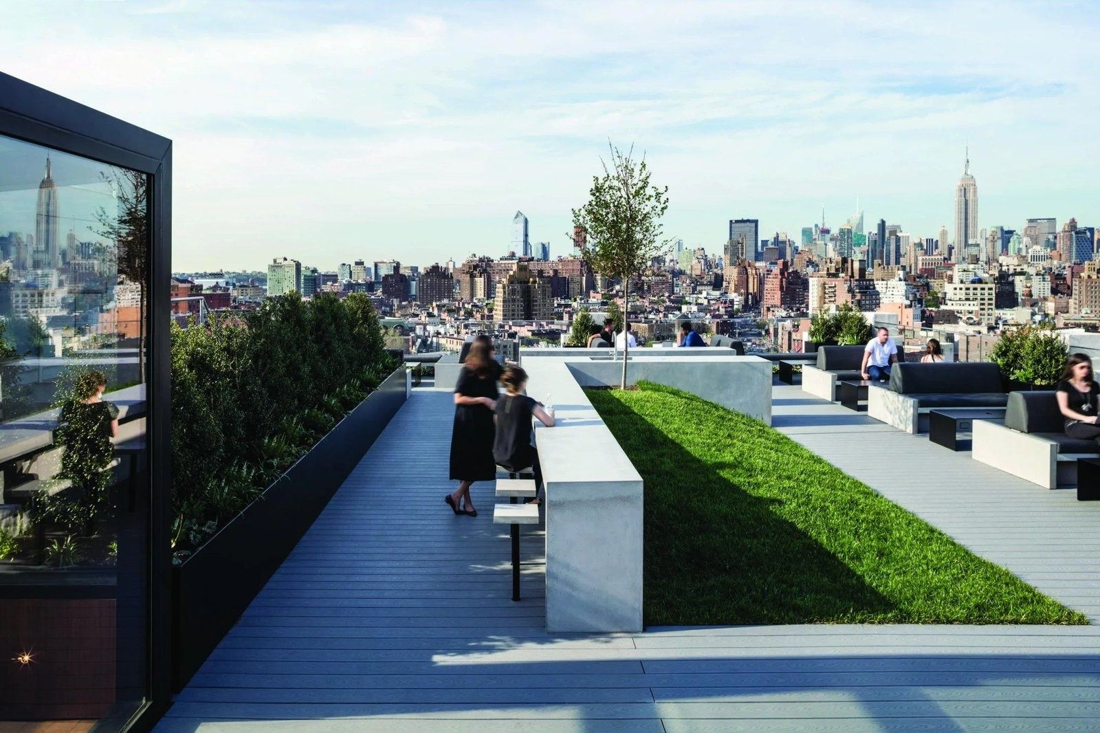 Rooftop deck with a strip of lawn, planters, concrete benches and a young tree, with the Midtown Manhattan skyline and the Empire State Building beyond