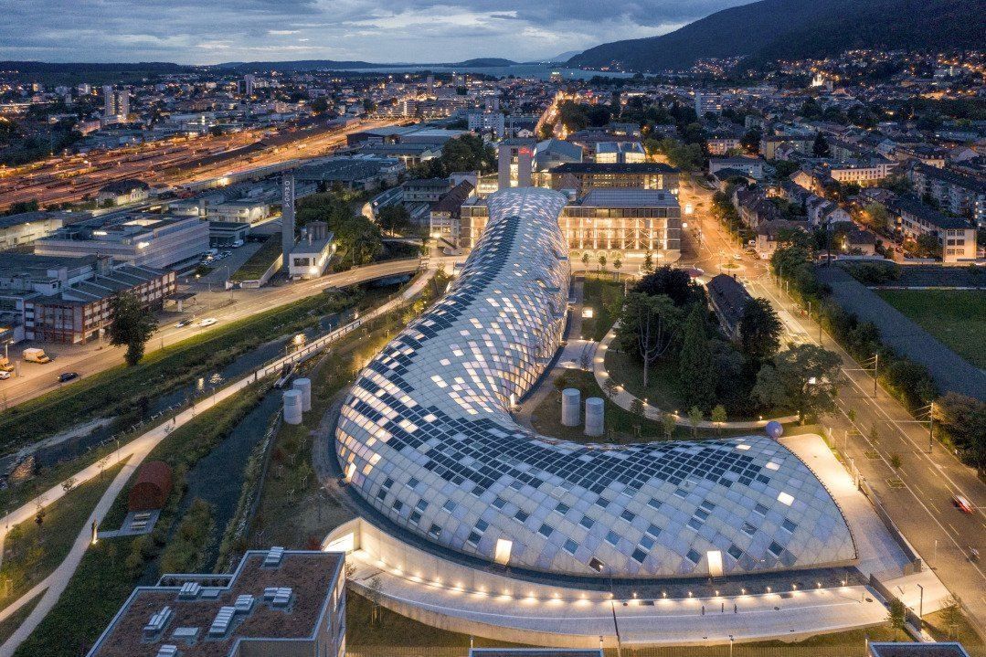 Aerial evening view of a long curved building with a white diamond-patterned roof beside a river and city lights