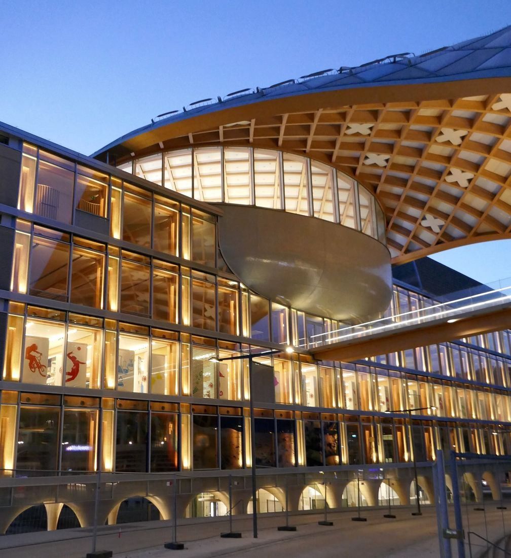 The Swatch Group: Glass building with arched ground floor and a silver oval under the timber canopy at dusk