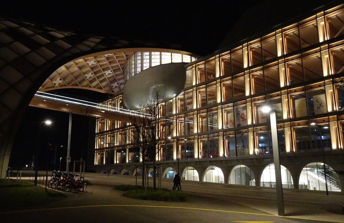 The Swatch Group: Night view of the illuminated glass building and the timber arch above a plaza