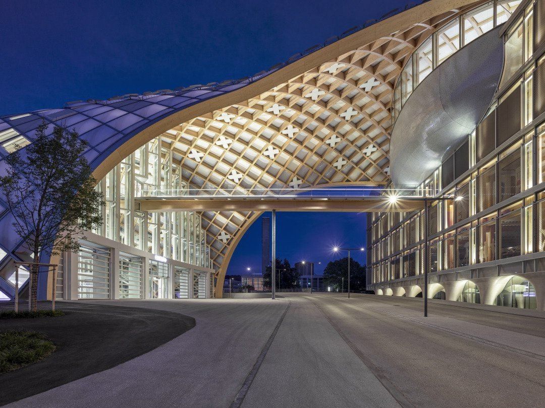 The Swatch Group: Timber grid arch spanning a street at dusk, with a silver oval form under it and a glass office building to the right