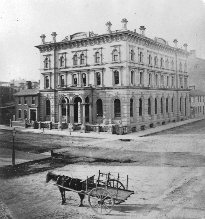 Black and white photo of an Italianate stone bank building on a corner, with a horse cart in front