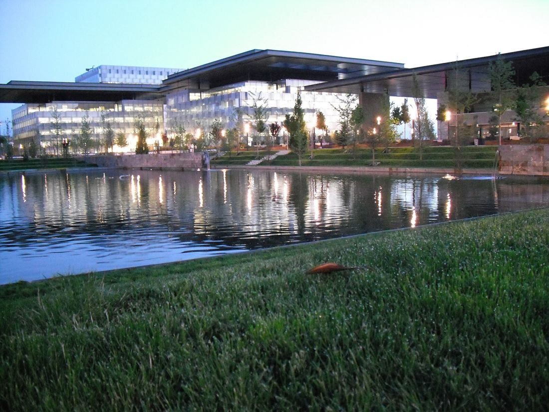 Low glass buildings under a flat canopy reflected in a lake at dusk, lawn in the foreground