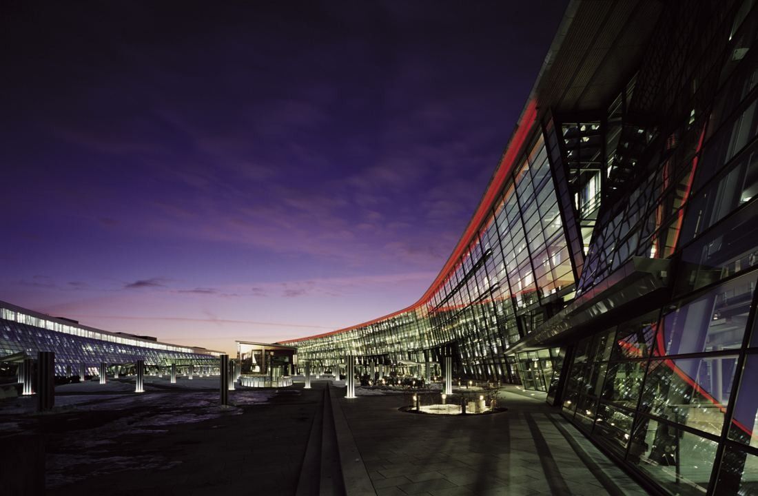 Curved glass facade with a red roof edge at dusk, with a wide paved square in front