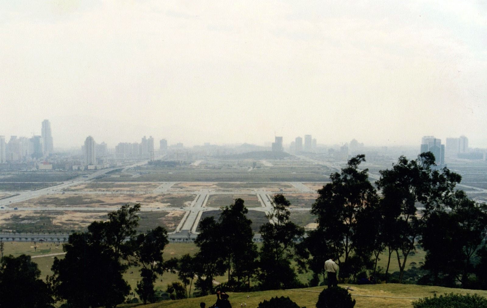 Hazy view over a flat, half-built city from a wooded hilltop