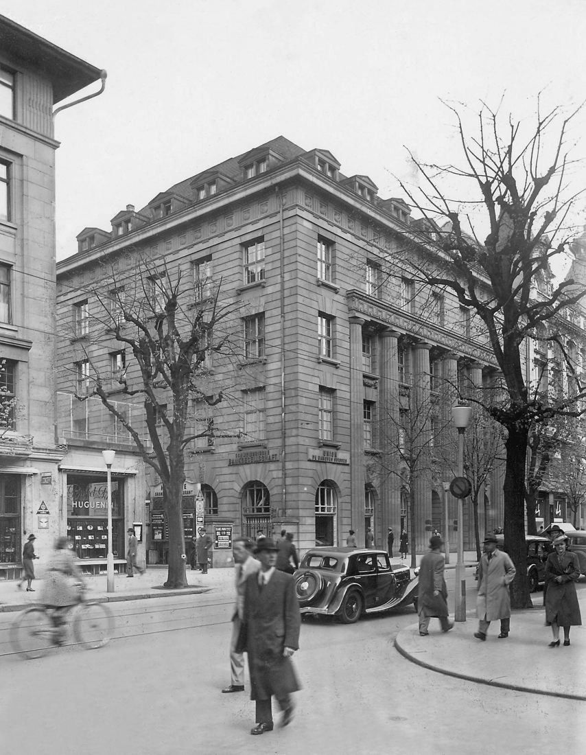Black-and-white photo of a large stone building on a street corner with bare trees and a few passers-by