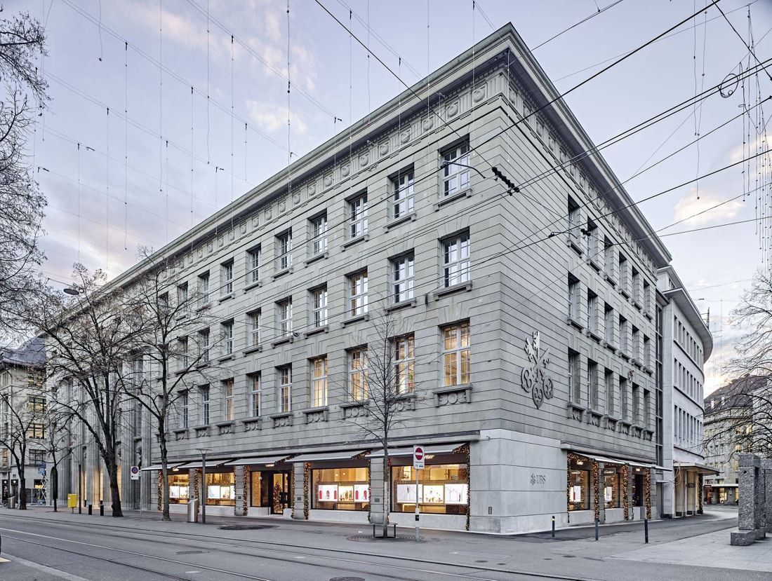 Corner view of a large grey stone bank building with lit shopfronts on the ground floor and tram wires above the street