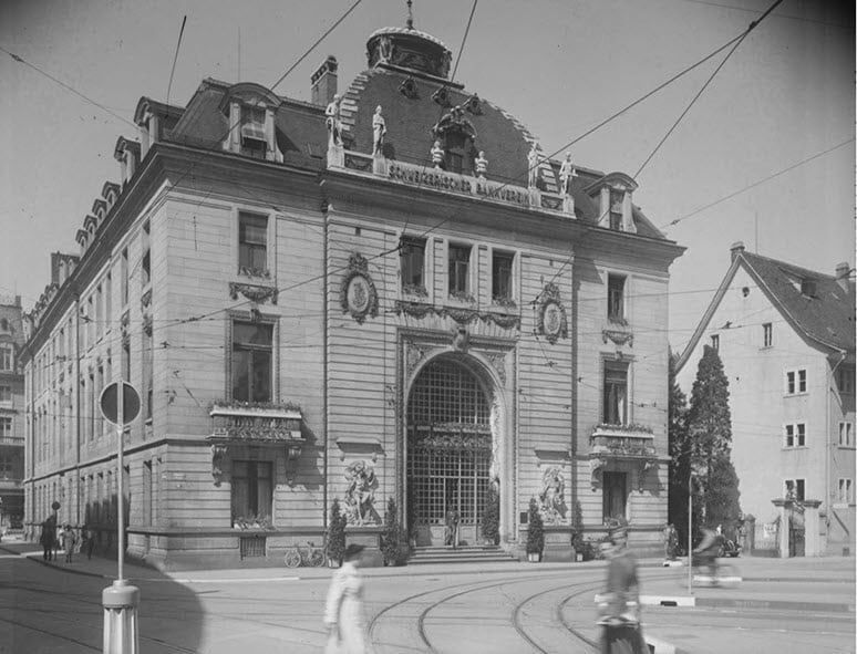 Stone bank building with a domed roof, statues and a large arched entrance, tram wires in front