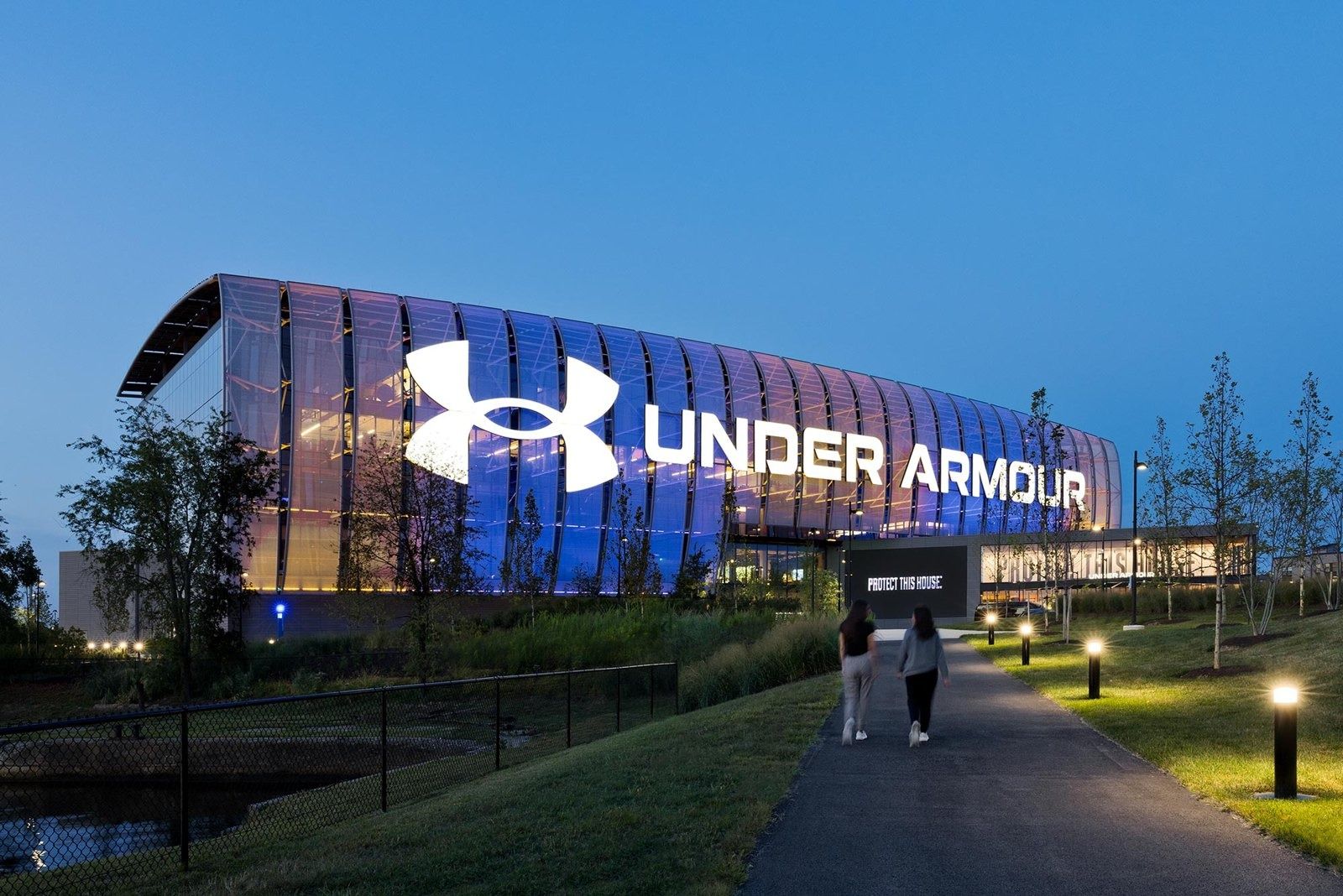 Curved glass building at dusk with a large illuminated Under Armour logo and lettering, a lit path in front