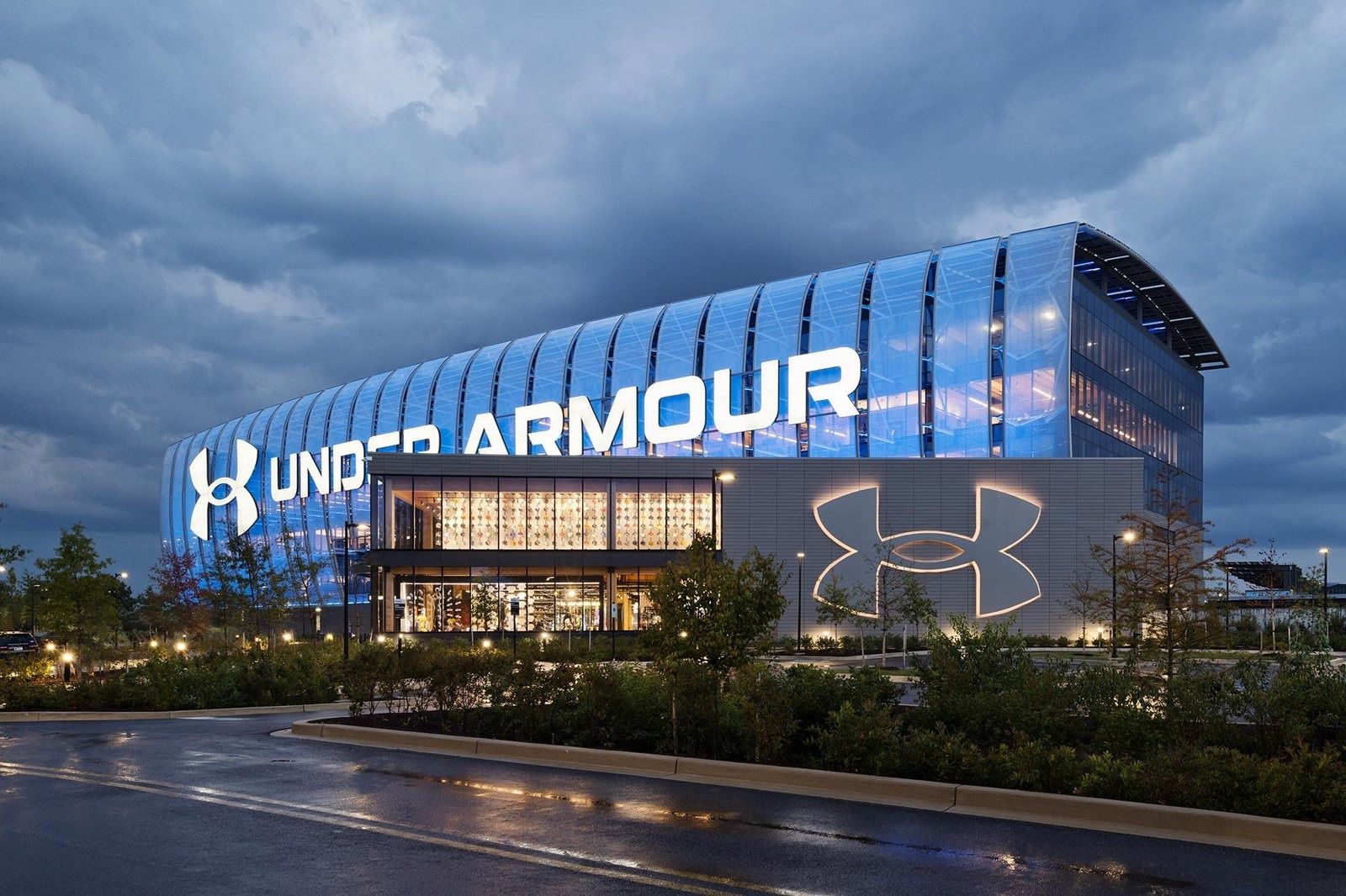 Under Armour: Glass building lit up at night with large Under Armour lettering and logo, wet road in the foreground