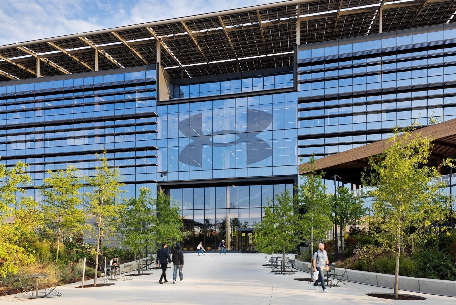 Under Armour: Glass office facade with a large logo and solar panels on the roof, a tree-lined plaza with people walking