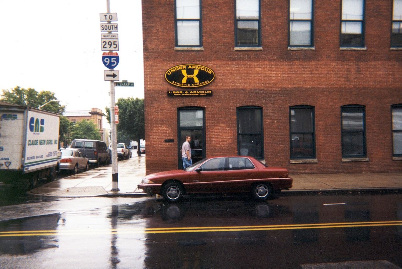 Red-brick building with an oval Under Armour sign, a parked red car and I-95 road signs on a wet street